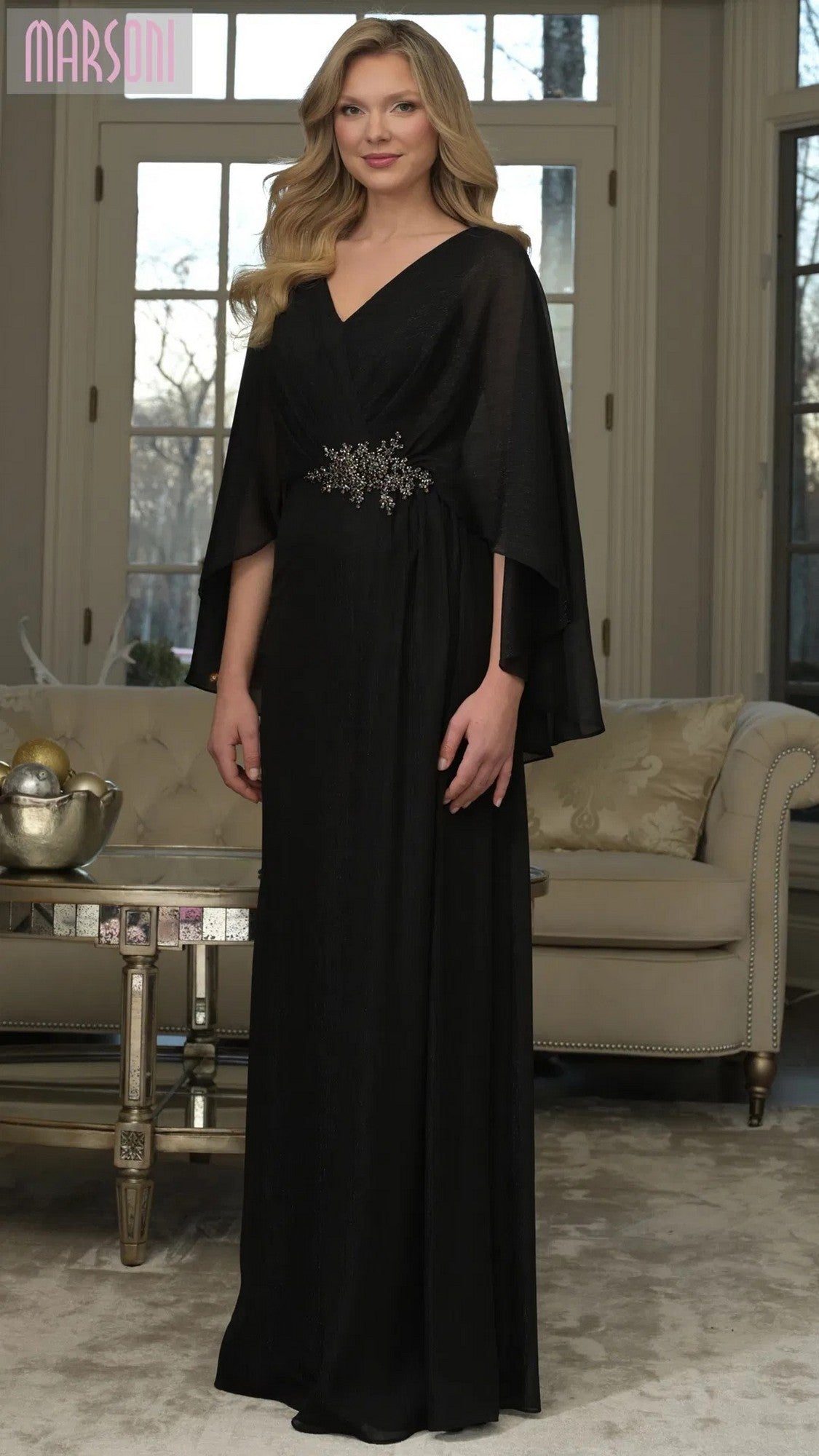 A woman wears the Marsoni MV1361 long formal dress in black, featuring cape sleeves and a crystal-beaded applique waistband, as she stands in a living room with a sofa and large windows behind her.
